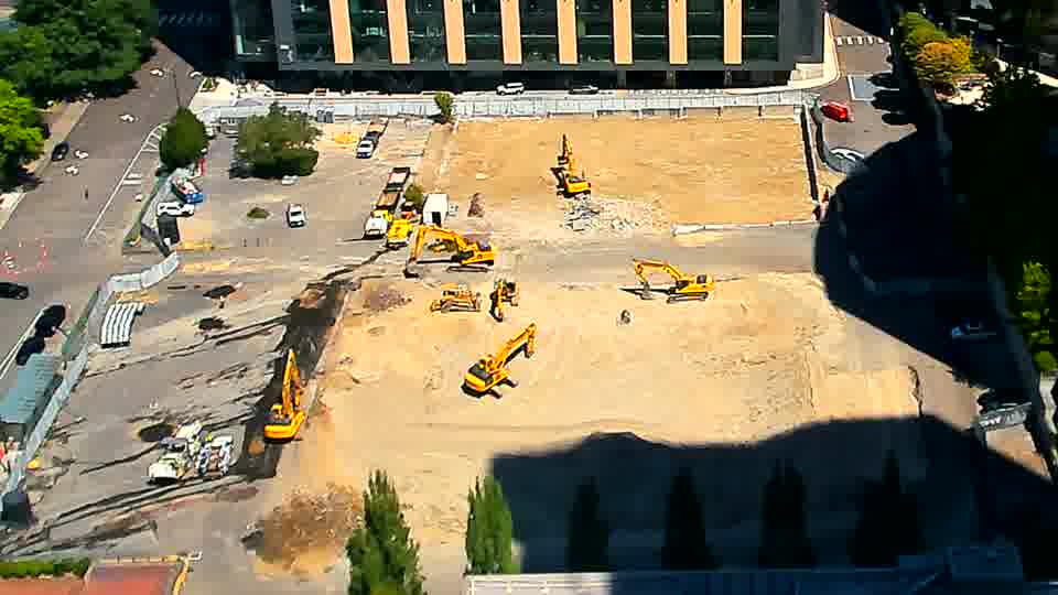 A light-colored truck is exiting the site via the driveway on the left, heading onto the public street. The positions of the larger machines and material piles appear to be holding steady.