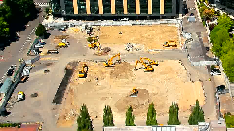 The machines on site are holding their positions, though a small white vehicle is now present near the trees at the far right.