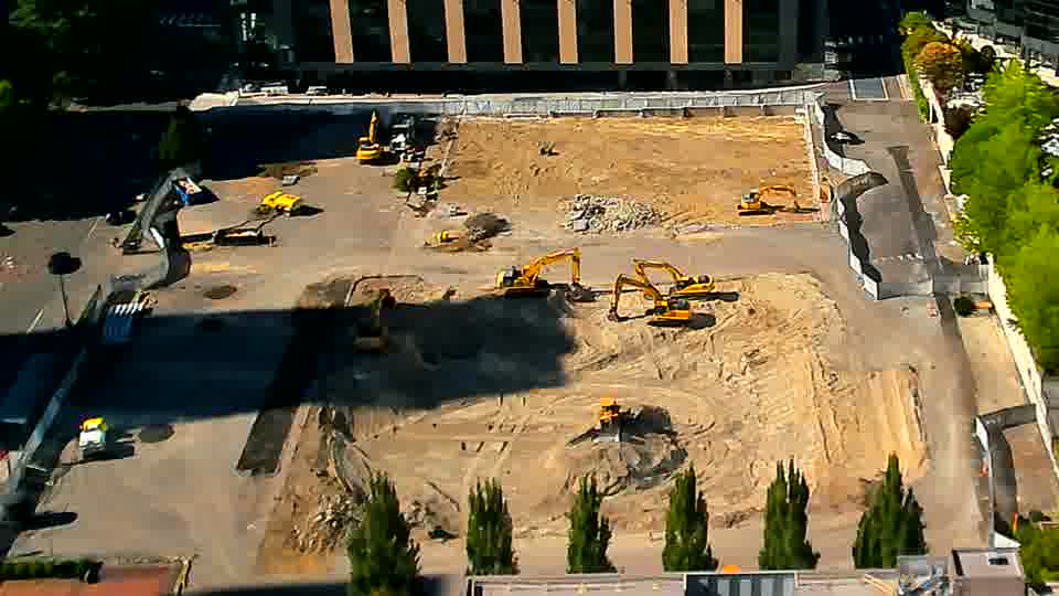 The layout of machines and materials is consistent between views, though a small white vehicle is now visible near the driveway on the left.
