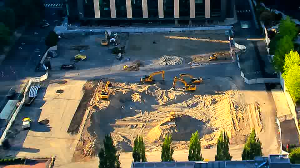 A small yellow vehicle has appeared near the driveway on the left side of the frame. Otherwise, the arrangement of equipment and materials across the site remains consistent.