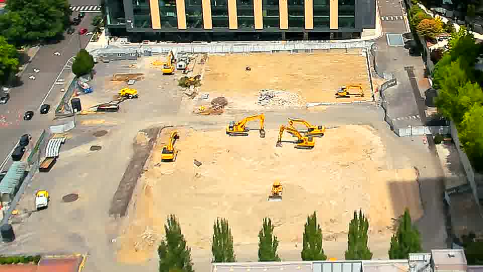 A small, light-colored vehicle has entered the site via the driveway on the left side of the frame. Otherwise, the equipment and general scene appear static between these views.