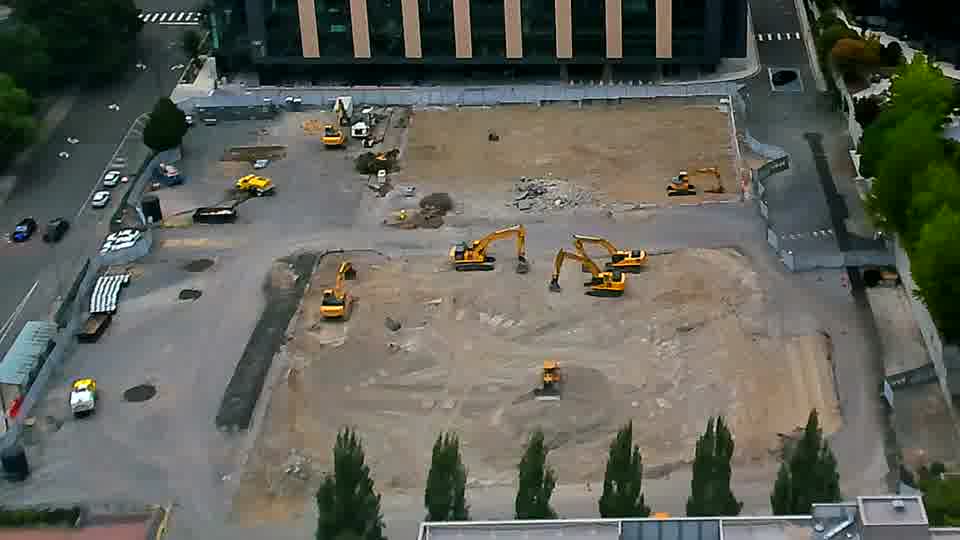 A white pickup truck has entered the site via the driveway on the left side of the frame. Otherwise, the equipment positions and overall scene appear consistent between the two views.