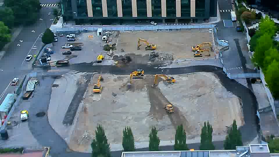 A yellow excavator near the center of the frame has shifted its position slightly, rotating to face a different direction. The rest of the site appears to be in the same condition as it was fifteen minutes ago.