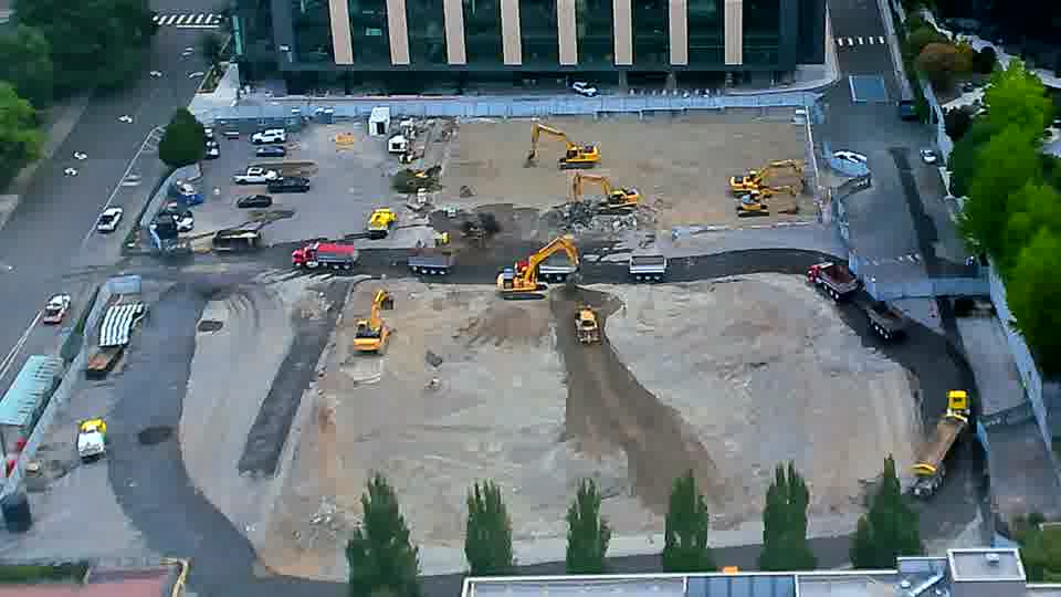 A red and black truck has entered the site from the driveway on the left, and another has moved further along the service road on the right. The arrangement of the larger equipment across the work area is otherwise similar to fifteen minutes ago.