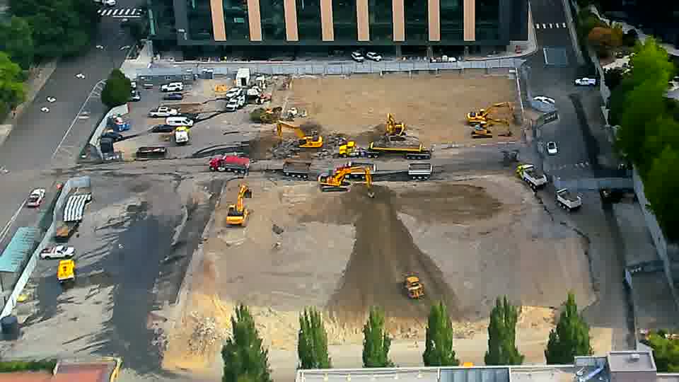 A few more vehicles are visible along the service road on the right side of the frame, but the heavy equipment remains in largely the same positions.