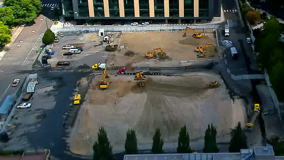 An excavator near the center of the site has moved a short distance toward the lower edge of the frame.