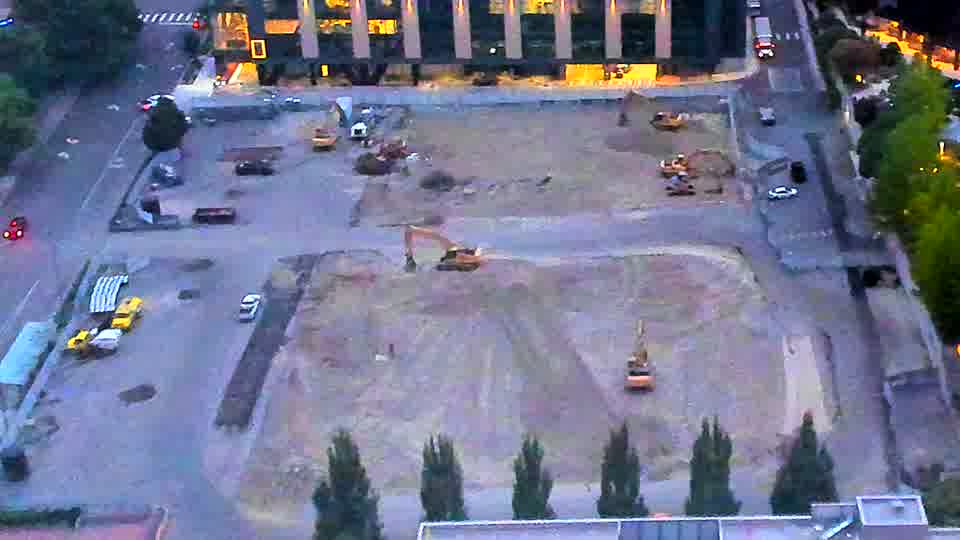 Daylight reveals several excavators and a dozer positioned across the site, with piles of material remaining near the northeast corner. The scene appears largely unchanged from fifteen minutes ago, with the same machines visible in similar locations.