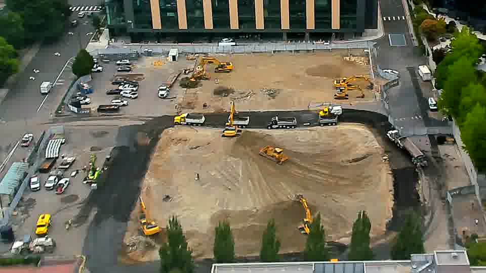A dump truck is now visible exiting the site via the driveway on the left, having moved from a position near the center of the frame. Otherwise, the arrangement of equipment appears consistent.