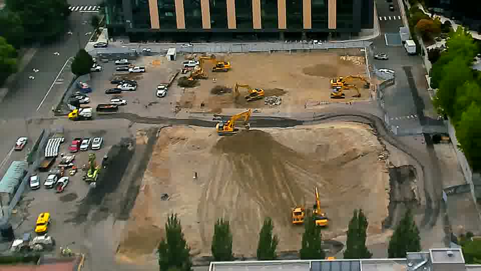 An excavator near the center of the site has lowered its arm, and its position appears slightly altered. Otherwise, the arrangement of equipment and vehicles remains largely as it was.