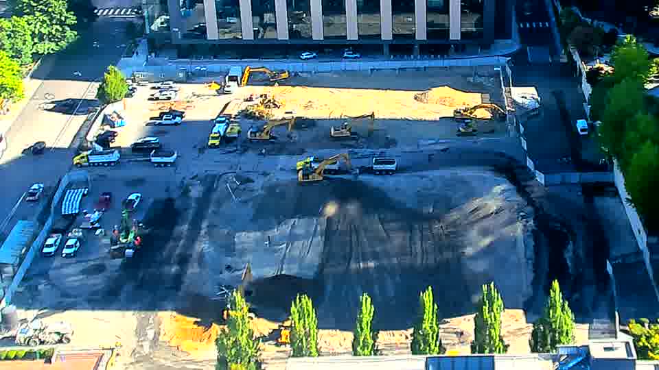 A light-colored truck has entered the driveway on the left side of the frame, and one of the excavators near the center has rotated slightly to the right.
