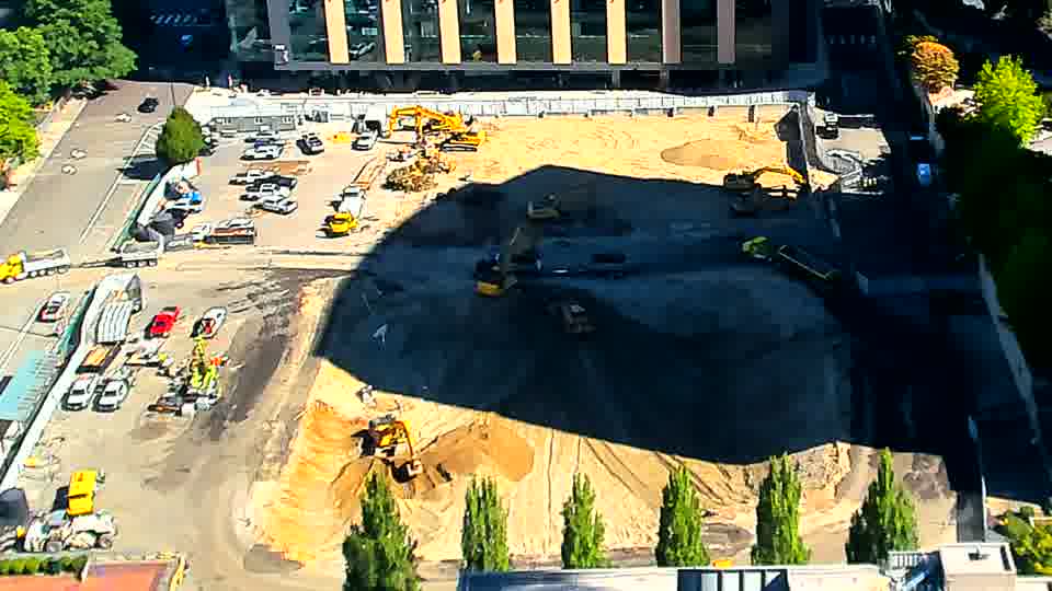 A handful of vehicles have gathered near the driveway on the left, and the excavator closest to the foreground appears to have shifted its orientation slightly.