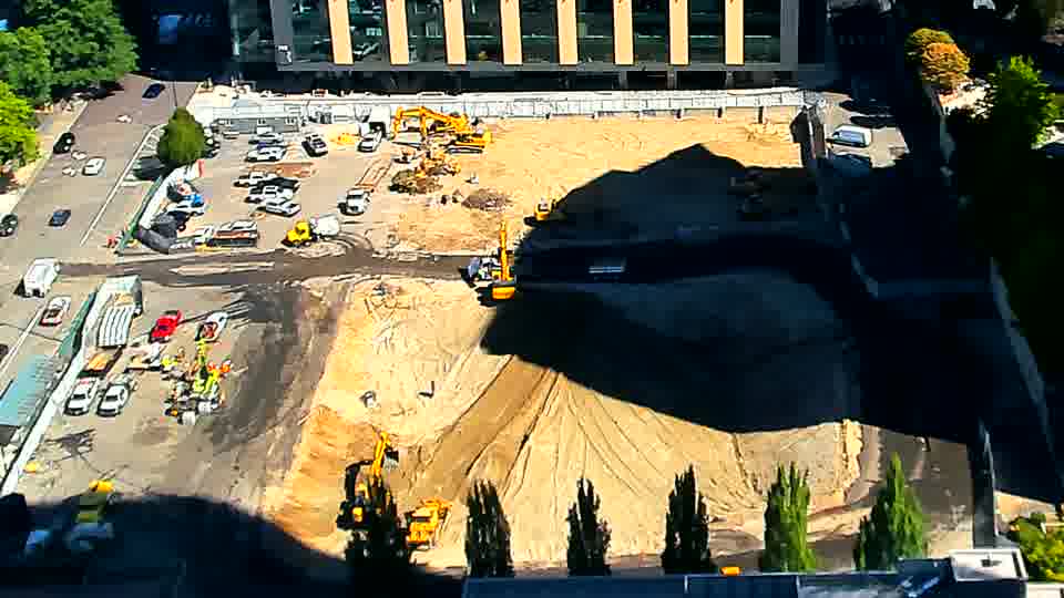 The excavator near the center of the frame has rotated slightly to the left, and a truck is now exiting the driveway on the left side of the image.