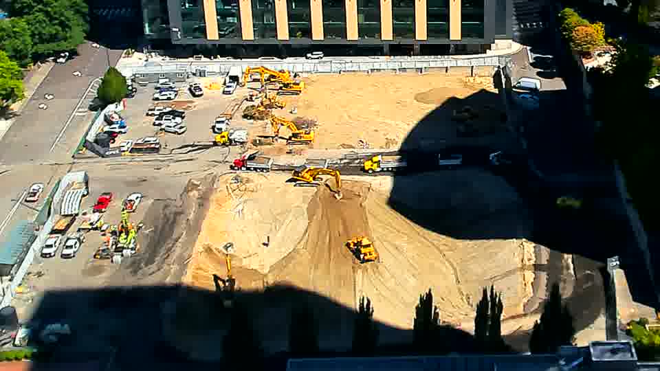 The excavator closest to the foreground is now positioned slightly further back from where it was, and a few additional vehicles are visible near the driveway on the left.
