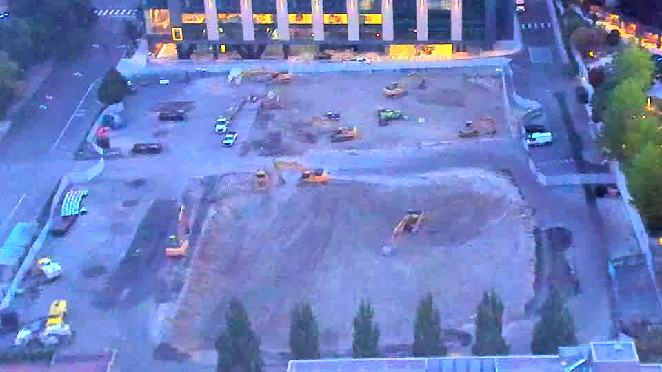 The site is now visible with increased light, revealing several pieces of heavy equipment scattered across the graded area. There is no apparent movement between the two frames, and the machines remain in the same positions.