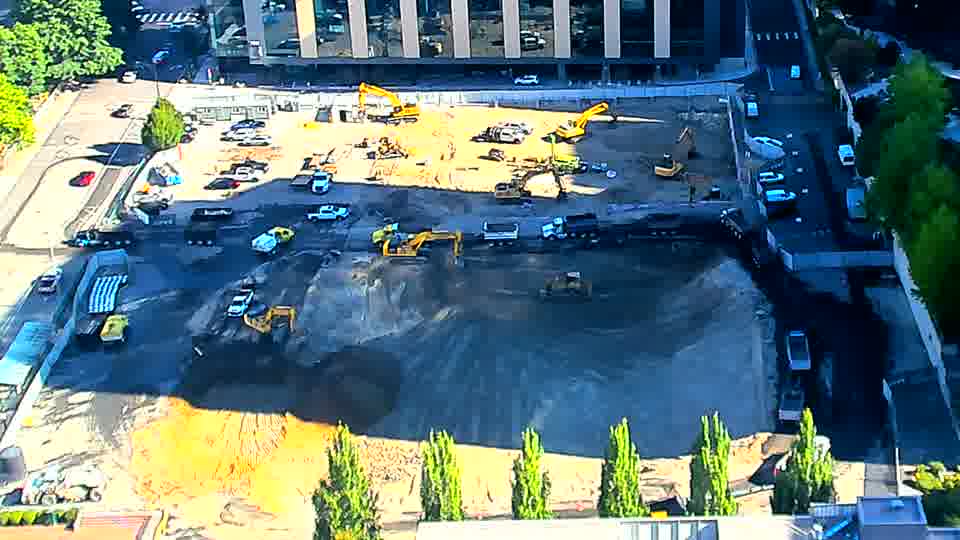 A small, blue vehicle has moved from the lower-left to the center of the site. Otherwise, the equipment and materials across the block remain in similar positions.