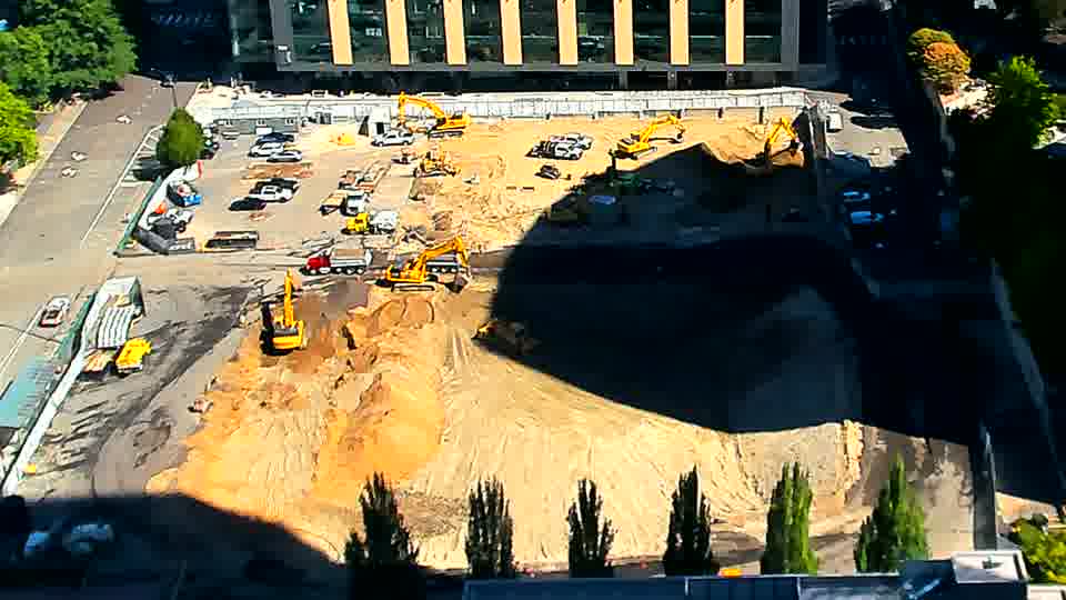 The layout of machines and materials appears nearly identical to the prior view, though the sunlight continues to shift across the site.