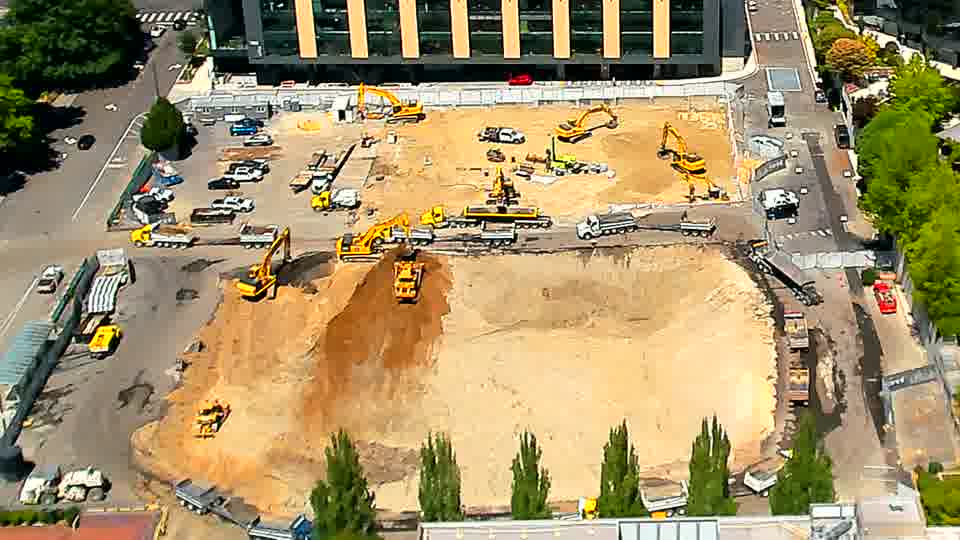 AnCSone excavator in the center of the site has shifted position, and a white truck has moved further along the central transit path. All other equipment and materials remain in place.