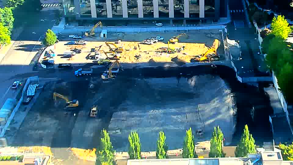 An excavator has moved from the center of the site to a position near the lower edge, and a second machine appears to have shifted slightly alongside it. The rest of the scene remains largely unchanged in terms of equipment placement.