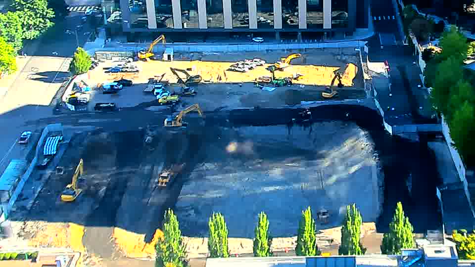 The excavator in the lower-left has shifted further toward the edge, and a blue truck near the center has moved. All other machinery and materials remainC remainLSS remain in place.