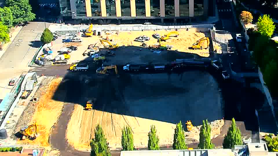A dark-colored vehicle has moved into the central area of the site, positioned between two of the larger yellow machines. Beyond that, the scene appears consistent with the previous view.