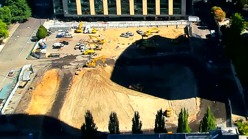 ALHKBS-A yellow laL-A yellow excavator in the center of the site has shifted position slightly, and a dark vehicle has departed from the central area. All other equipment and materials remain in place.