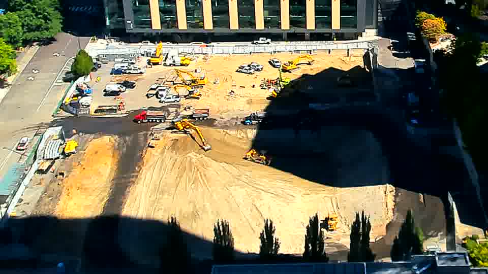 ABL-A red dumpB-A red dump truck has entered the site and is stopped near the center, while a yellow excavator in that same area has shifted its position. No other significant changes are visible across the block.