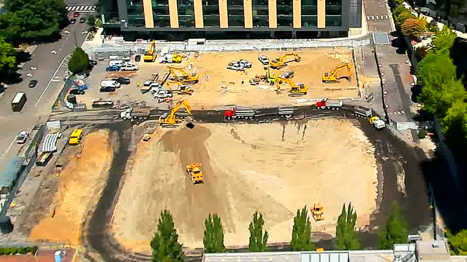 An excavator near the lower-right corner of the site is now facing a different direction. Otherwise, the equipment and materials across the block appear to be in the same locations as before.