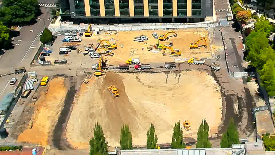 A small dump truck has entered the site from the driveway on the left, then moved toward the center. Otherwise, the equipment and materials maintain their previous positions across the block.
