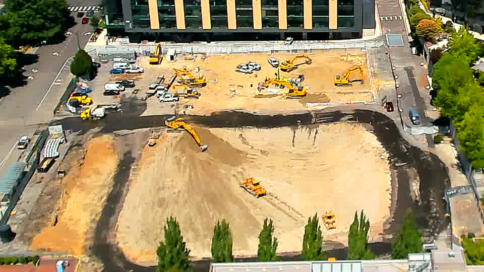 An excavator in the lower center of the site is now actively moving material, having shifted its boom and bucket. The rest of the equipment and stockpiled materials remain in largely the same arrangement as fifteen minutes ago.