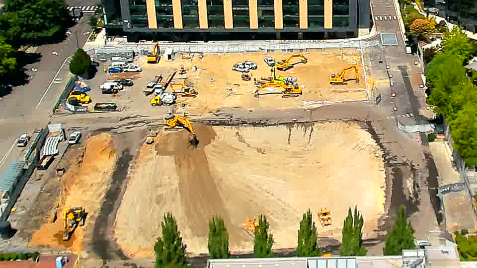 The large excavator in the lower center of the site now has a different angle to its boom and bucket. Otherwise, the arrangement of equipment and materials across the site appears largely unchanged.