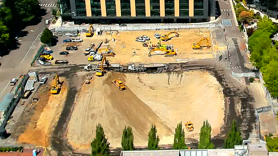 The position of the small, yellow machine in the lower-center of the site has changed, now facing a slightly different direction. The rest of the equipment and materials across the block appear to be holding steady.