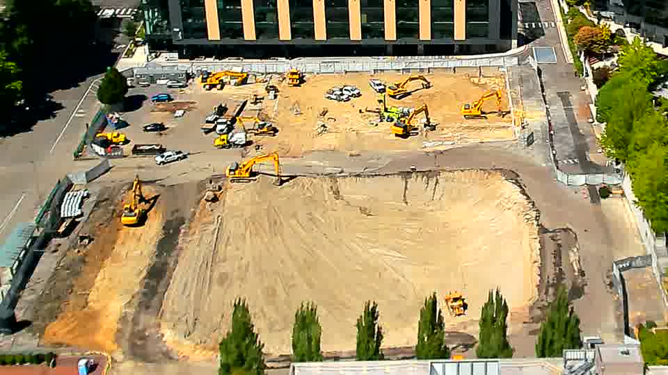 A small, white pickup truck has entered the site via the driveway on the left side of the frame. Beyond that, the equipment and material arrangements on the block are consistent with the previous view.