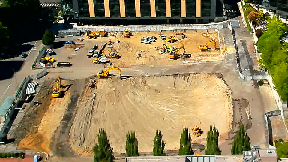 A white vehicle is now visible near the center of the site, having appeared between views. Otherwise, the arrangement of heavy equipment and graded areas remains consistent across the block.