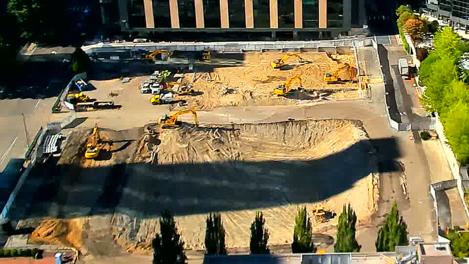 A few figures are now visible near the center of the site, clustered around one of the excavators. Otherwise, the layout of the site and equipment remains largely as it was.