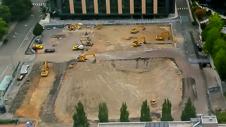 The site appears largely static, though a small yellow machine has become visible near the service road on the right side.