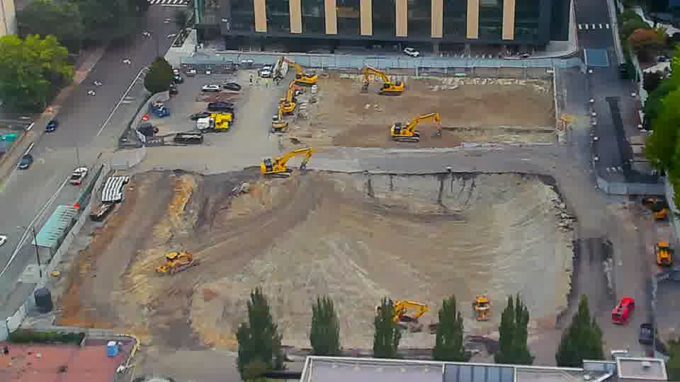 A small red vehicle has appeared near the equipment in the southeast corner of the site. Beyond that, the positioning of machines and materials looks the same as it did moments ago.