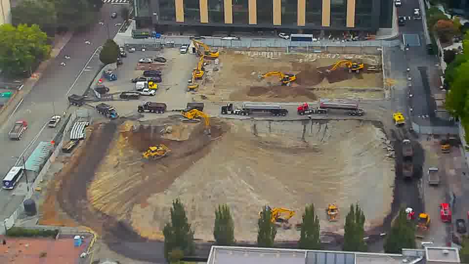Several trucks and a dark-colored loader are now visible along the service road on the right side of the frame, having appeared since the last view.