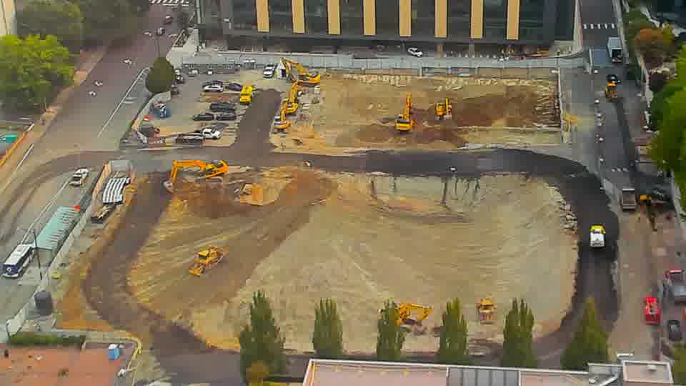 A white box truck has entered the site from the driveway on the left side of the frame and is now positioned near the center. Otherwise, the arrangement of equipment and materials remains consistent.