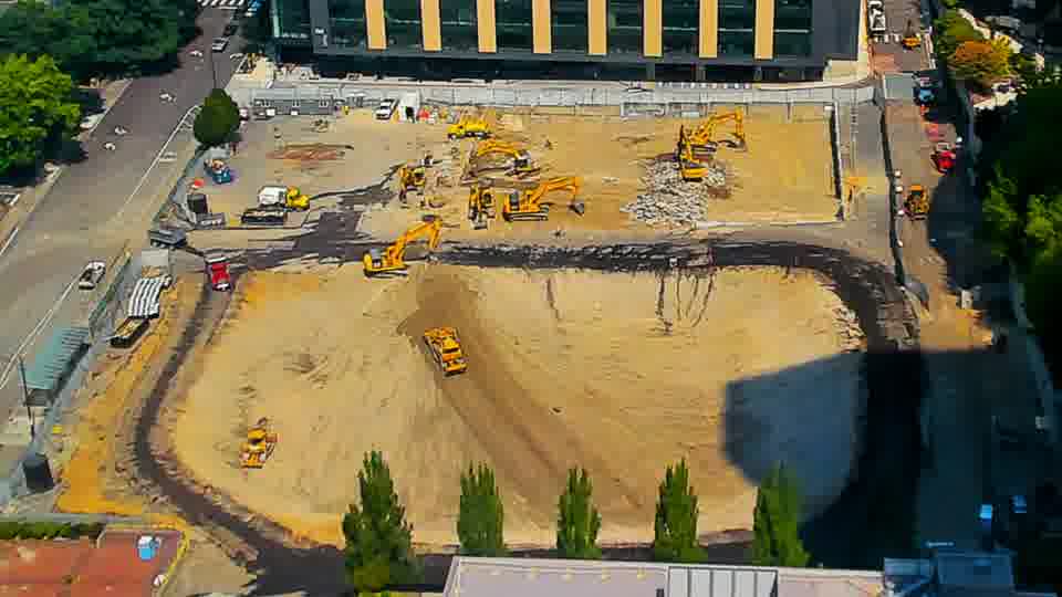 A yellow machine near the center has advanced slightly further into the graded area. The overall layout of equipment and materials remains otherwise stable.