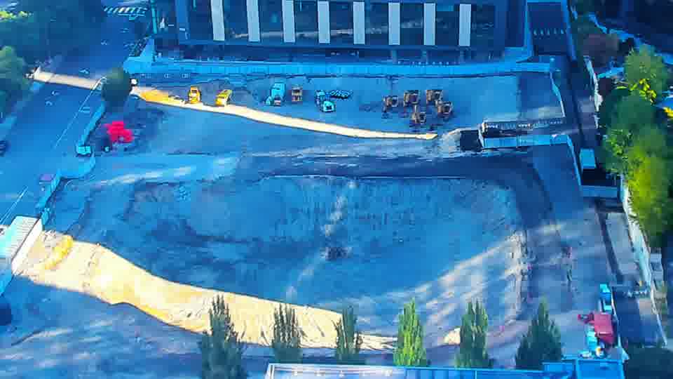 Construction camera frame at 9:00 AM PT. A yellow machine has shifted position within the central area of the site. Otherwise, the scene remains consistent, with no other visible changes to equipment or vehicle locations.