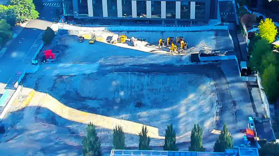 Construction camera frame at 9:15 AM PT. The arrangement of machinery in the center of the site has subtly altered, with one yellow piece of equipment appearing to have rotated. Otherwise, the scene presents a familiar picture, with no other notable shifts in equipment or vehicle positions.