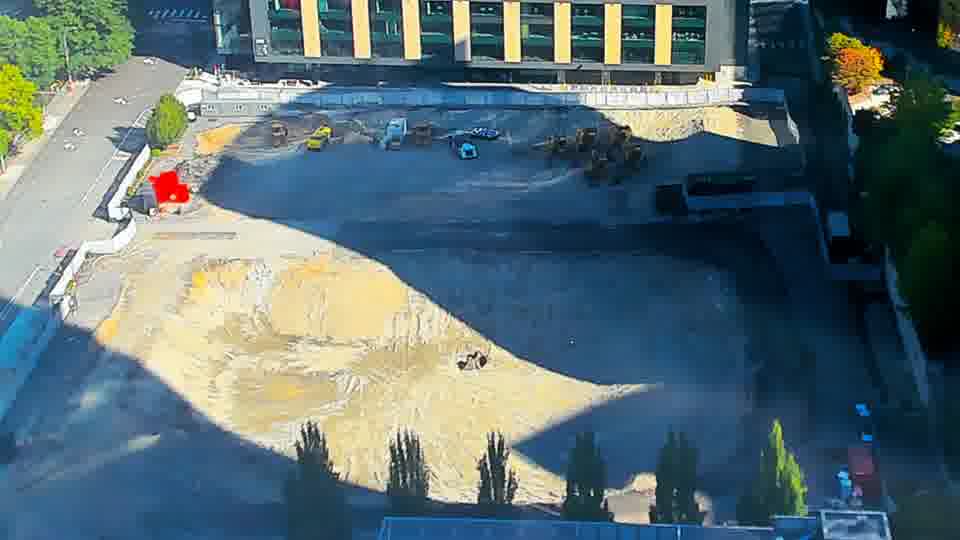 Construction camera frame at 10:15 AM PT. A small red structure has appeared near the center of the site, adjacent to the large pile of earth-toned material. The rest of the scene remains unchanged, with equipment and vehicles holding their prior positions.
