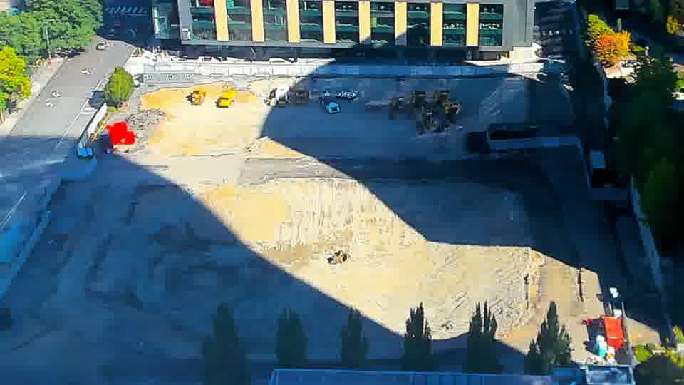 Construction camera frame at 10:45 AM PT. A small, light-blue machine is now visible among the other equipment near the center of the site; it wasn't present in the earlier view. The rest of the scene, including the red structure and the arrangement of materials, appears constant.