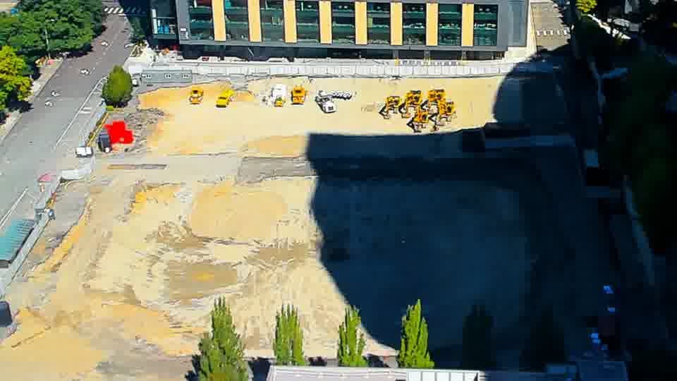 Construction camera frame at 12:00 PM PT. A white vehicle is now entering the site via the driveway on the left side of the frame. Beyond that, the arrangement of equipment and materials appears unchanged.