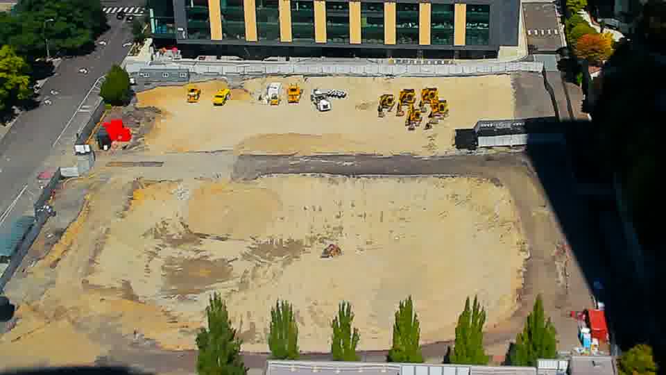 Construction camera frame at 12:45 PM PT. The arrangement of machines and materials across the site appears consistent between these frames, though shadows have continued to lengthen.