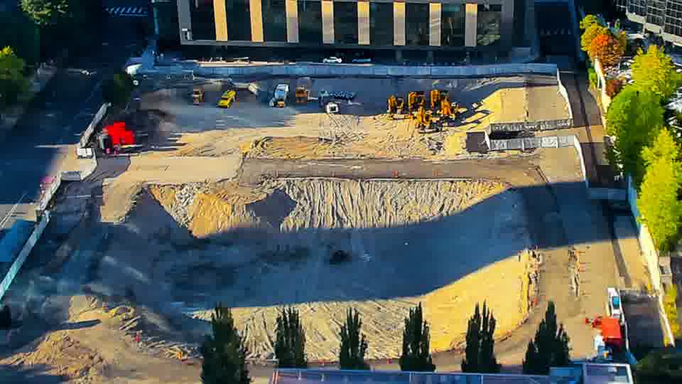 Construction camera frame at 6:00 PM PT. A white pickup truck has entered the site via the driveway on the left side of the frame. Otherwise, the arrangement of equipment and materials appears unchanged from fifteen minutes ago.