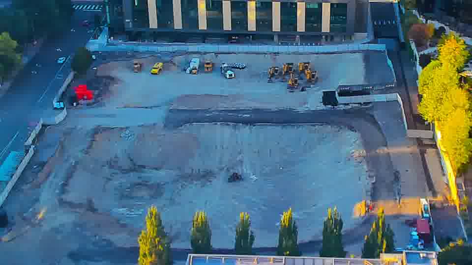 Construction camera frame at 7:00 PM PT. A small, dark-colored machine has moved from the right side of the central graded area toward the lower-center of the frame. Otherwise, the equipment and materials on site hold their positions.