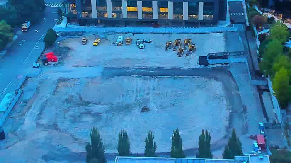 Construction camera frame at 7:15 AM PT. A white pickup truck has entered the site via the driveway on the left, and is now parked near the equipment cluster. Otherwise, the arrangement of machines across the graded area remains consistent between the two views.