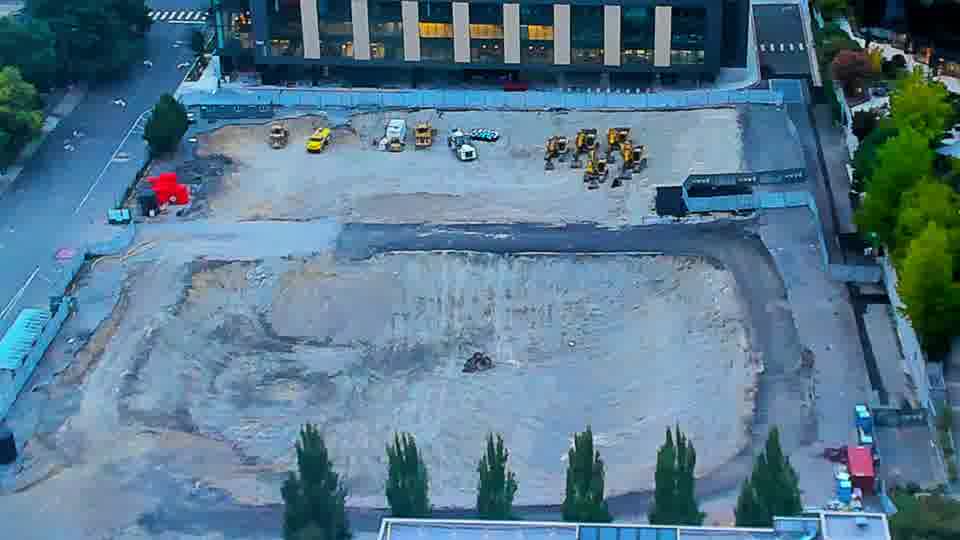 Construction camera frame at 7:30 AM PT. A small red vehicle has moved from the street-side edge of the site into the work area. The arrangement of larger equipment across the graded space appears otherwise unchanged.