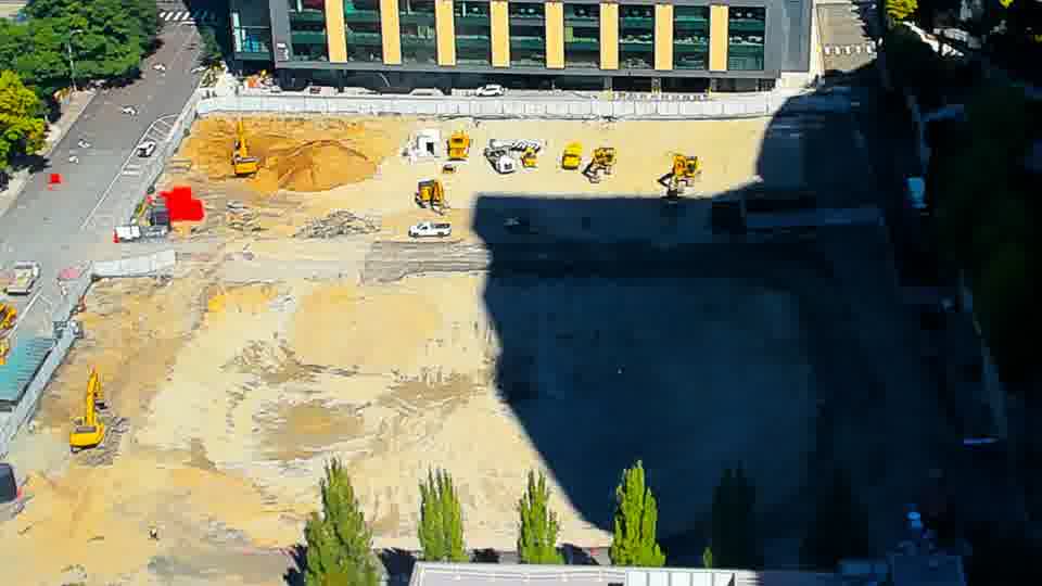 Construction camera frame at 12:00 PM PT. A small white vehicle is now visible in the central area of the site, having appeared between these frames.